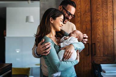 Une jeune mère et un père souriants tenant leur nouveau-né dans leurs bras à la maison. La scène chaleureuse et lumineuse illustre la joie de la famille, la parentalité et le lien affectif entre les parents et leur bébé.
